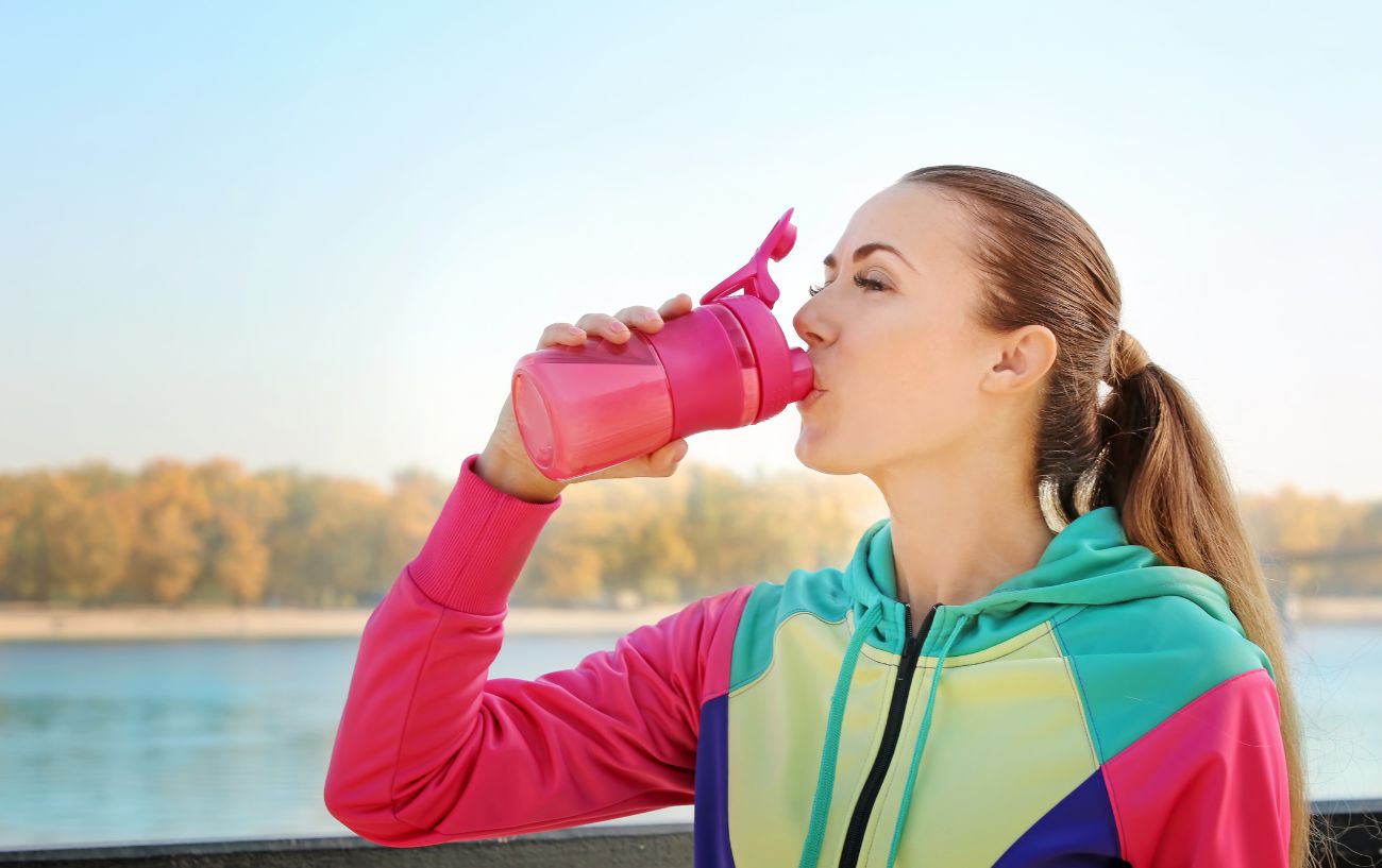 A person in a colorful sweatshirt drinking from a pink protein shaker.